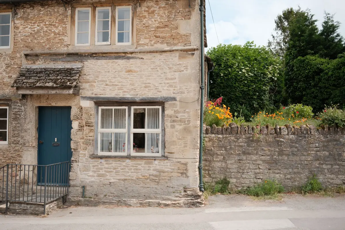 A stone cottage with a blue front door and a dry-stone garden wall on a village street in Lacock