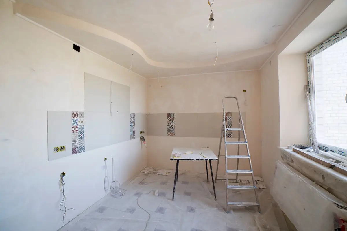 A kitchen mid-renovation: fresh plaster, new sockets, a stepladder and a trestle table on a protected floor