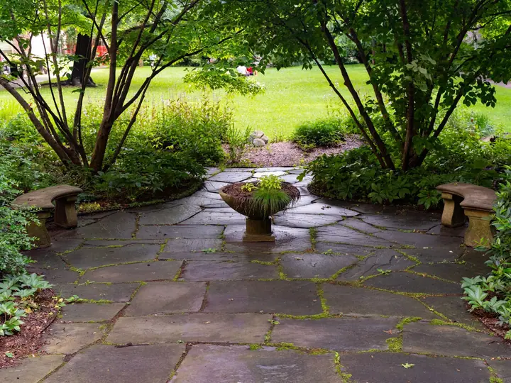 A flagstone patio with two stone benches, set between shrubs at the edge of a lawn