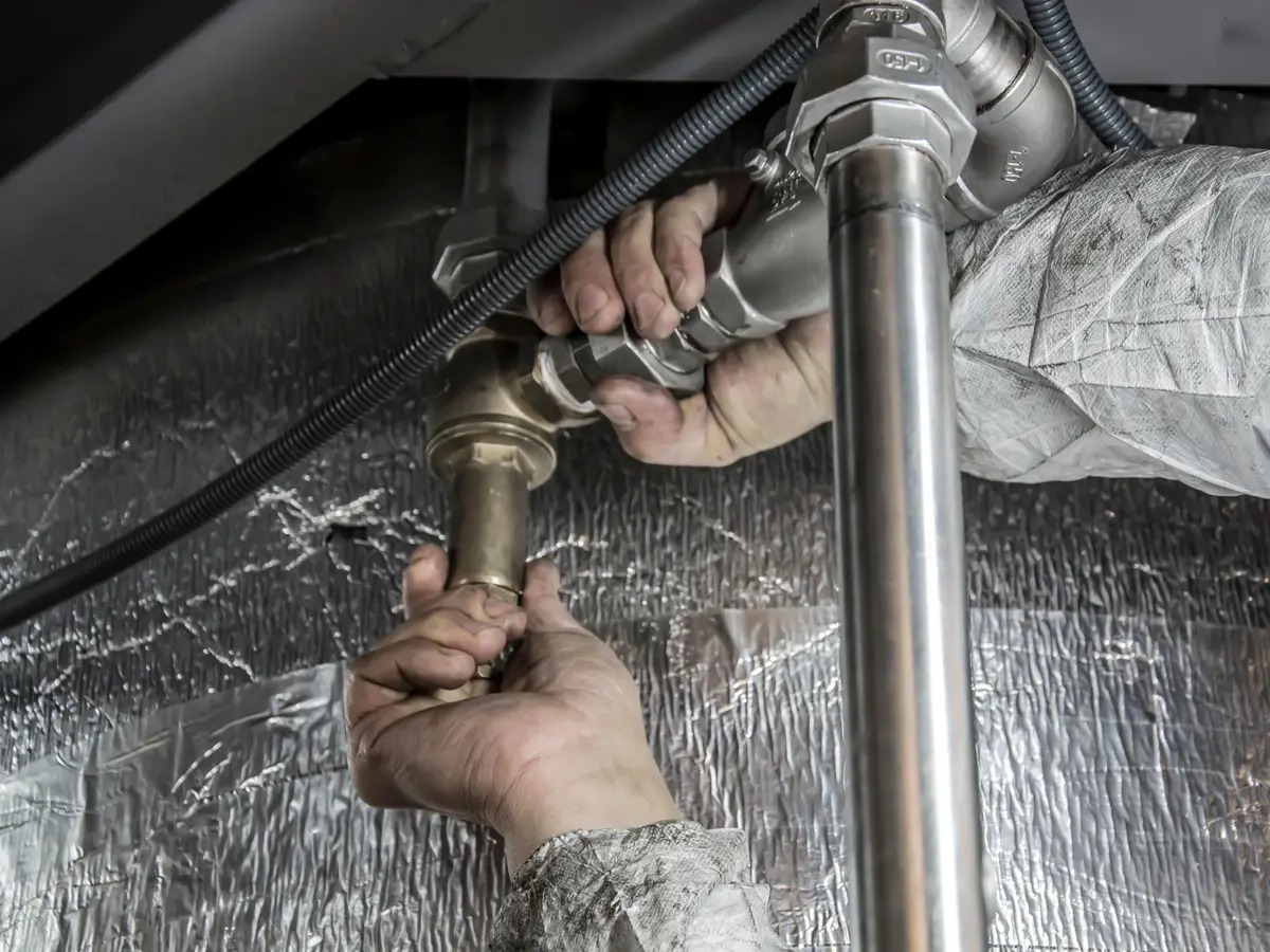 A plumber's hands fitting a brass valve onto a copper pipe beside foil insulation