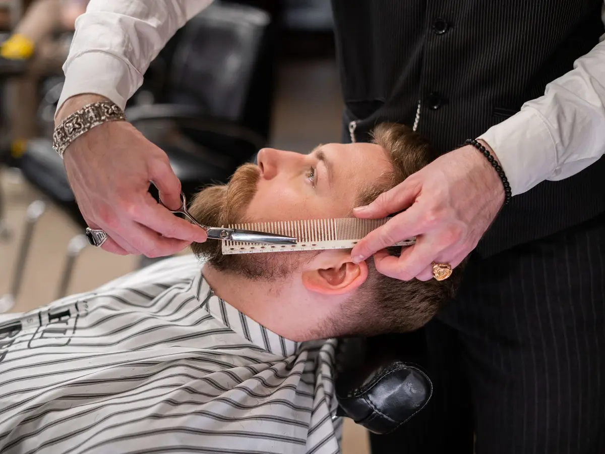 A barber trimming a client's beard with scissors and a comb
