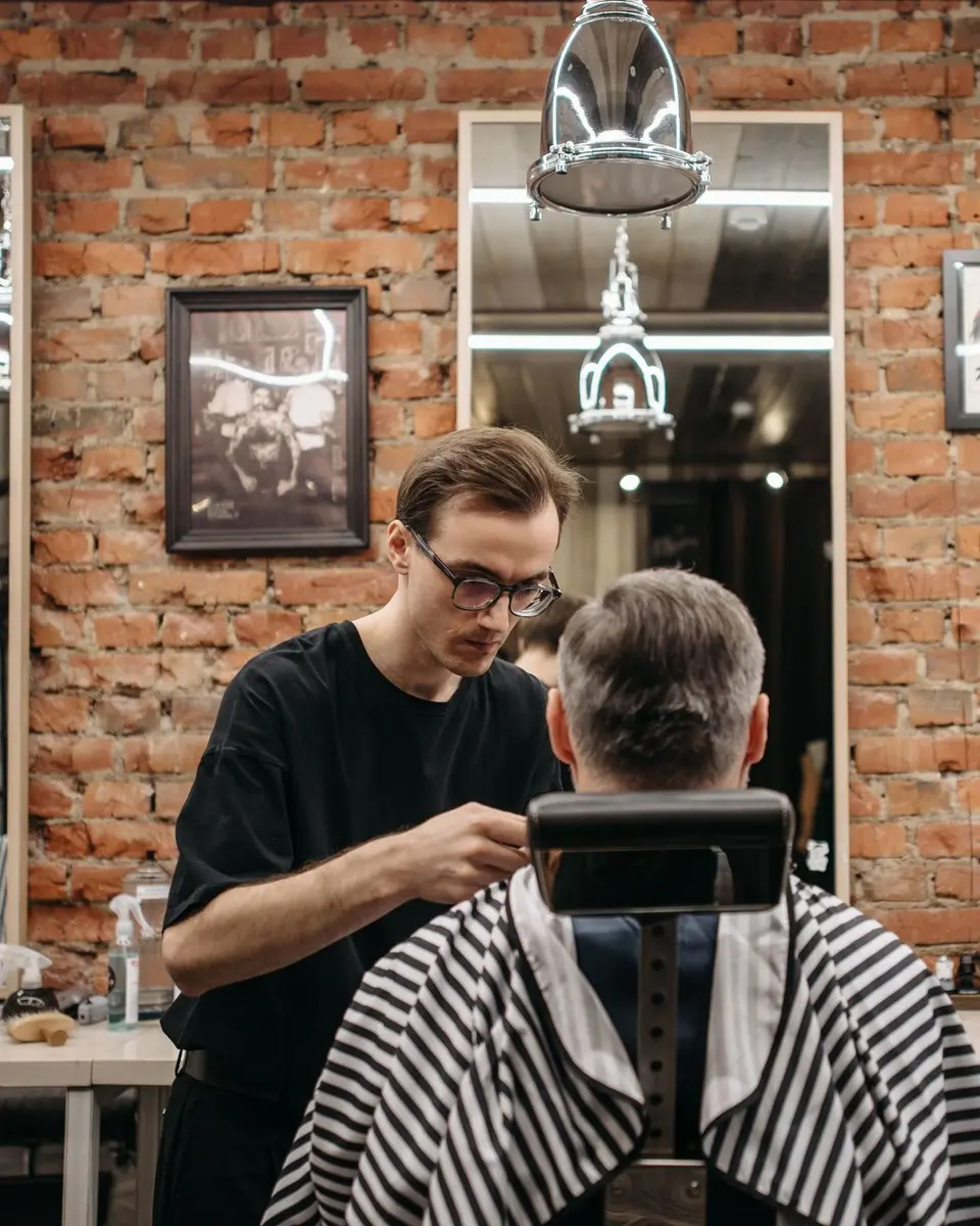 A barber in glasses cutting a client's hair in front of a bare red-brick wall, the client in a striped cape
