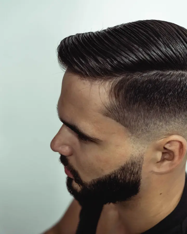 A man in profile with a slicked side part and a short, shaped beard