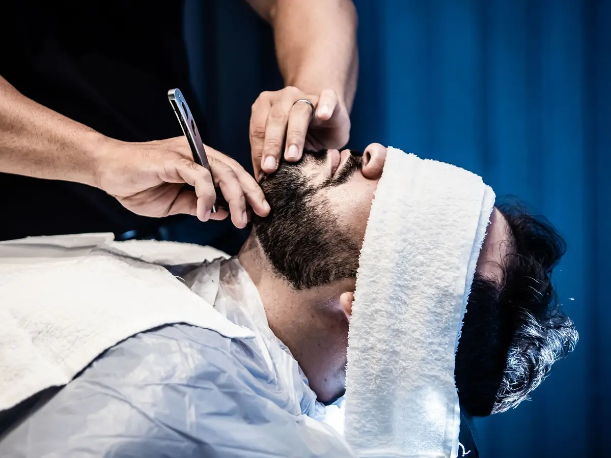 A barber shaping a reclined client's beard with a straight razor, a towel across his forehead