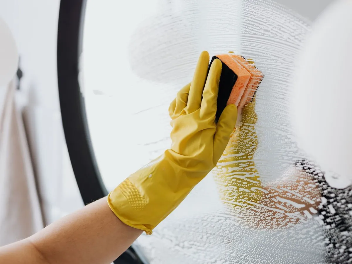 A yellow-gloved hand scrubbing a soapy bathroom mirror with a sponge