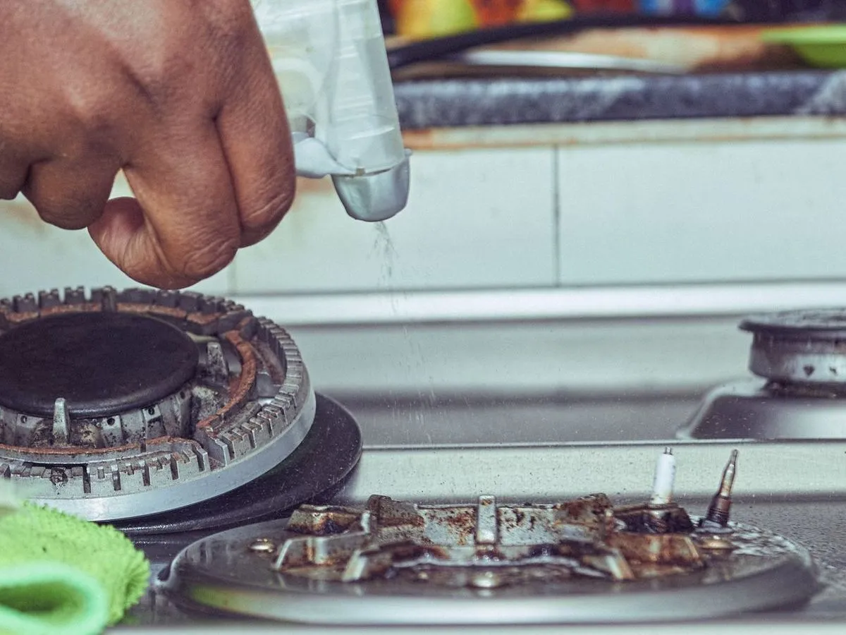 A hand spraying cleaner onto the burners of a gas hob, with a green cloth beside it