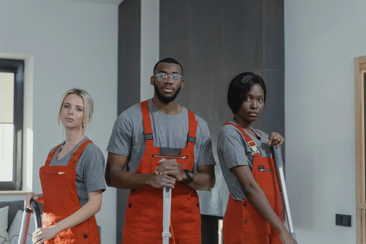 Three cleaners in matching orange overalls and grey T-shirts standing in a living room